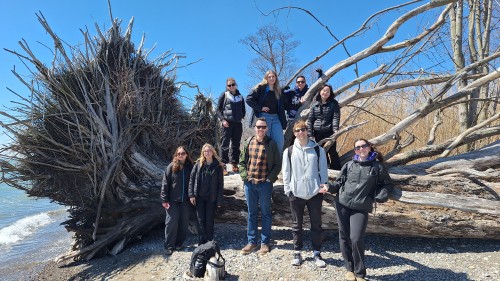 a group of students standing in front of a massive, felled tree on the ground on a waterfront