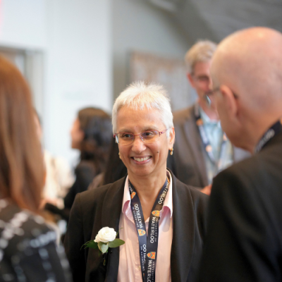 A smiling woman wearing glasses and a corsage, engaged in conversation with a woman and man at a formal gathering.
