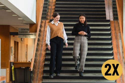 two students walking down the stairs in Tatham Center with a yellow circle on it with the text '2X' in it