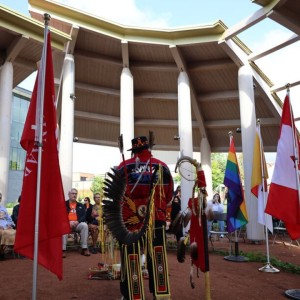 An Indigenous Elder wearing traditional regalia standing under a pavilion, facing people seated in a circle, surrounded by national and Pride flags.