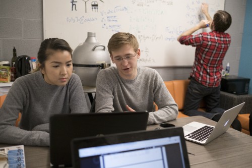 Two students looking at a laptop while another student works at a whiteboard