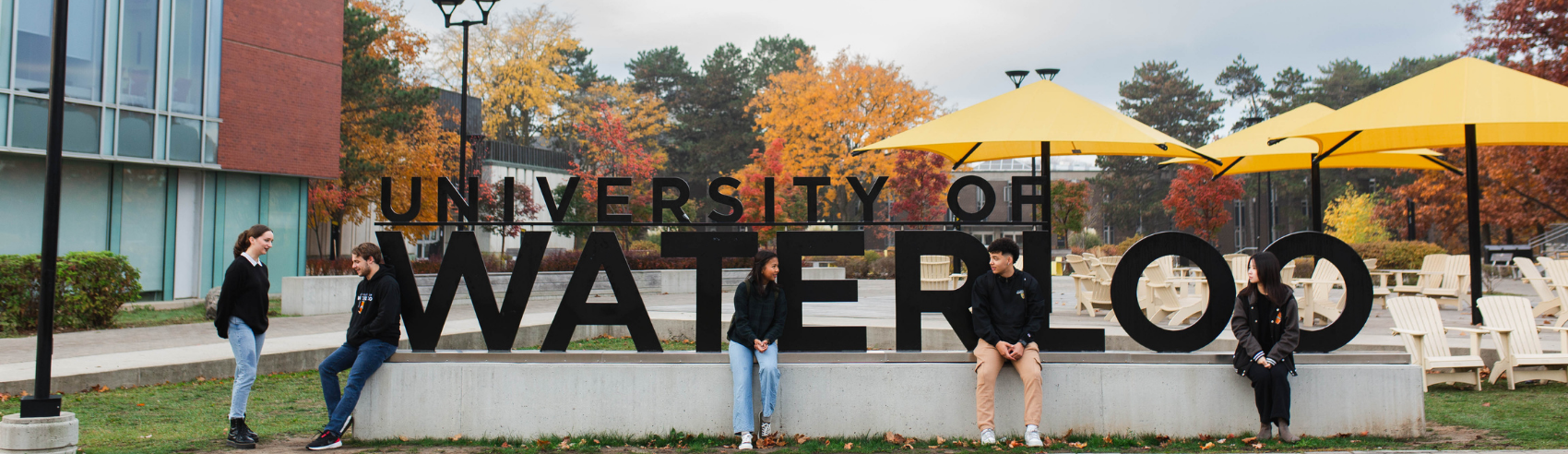 five students standing and sitting on the plinth holding a large 'University of Waterloo' sign on the Waterloo campus