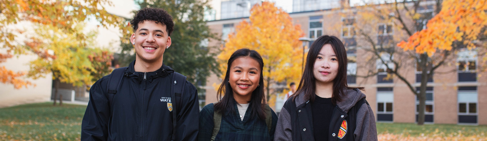 Three students standing and smiling in front of autumnal trees on the Waterloo campus